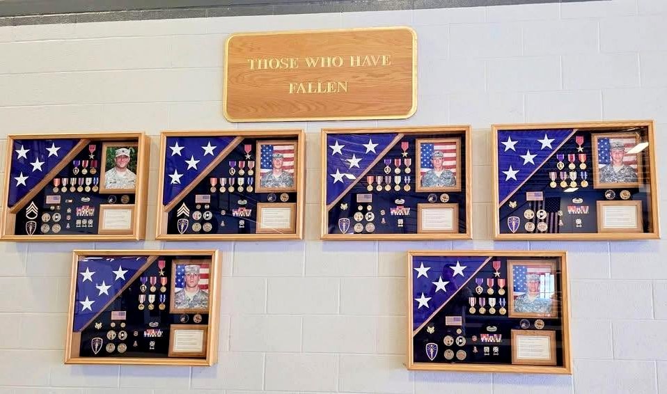 Six shadow boxes mounted on the armory wall of the 713th Engineer Company beneath a wooden plaque reading THOSE WHO HAVE FALLEN. Each box holds a folded flag, medals, unit coins and a photograph of the soldier.